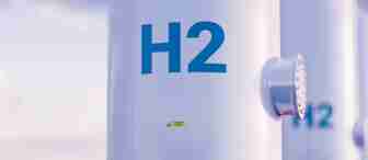 Close-up of a white cylindrical tank with "H2" written in blue, indicating hydrogen storage. The tank has a small valve or connector on the side. The background is slightly blurred with a light pinkish hue sky.