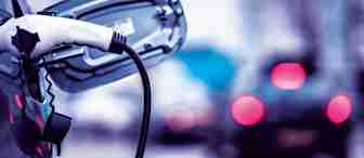 A close-up of an electric vehicle being charged, with the charging cable plugged into the car's port. The background features out-of-focus cars driving on a city street, creating a blurred effect with red taillights.