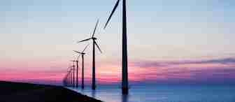 A row of wind turbines stands tall against a vibrant sunset backdrop over a body of water. The sky is painted with hues of pink, purple, and blue as the day transitions into evening. The turbines are aligned in perspective, stretching into the distance.