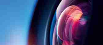 Close-up shot of a camera lens reflecting colorful light. The lens exhibits a spectrum of colors including pink, purple, and blue, with a blurred, gradient blue background that transitions from dark to light. The image emphasizes the intricate details of the lens.