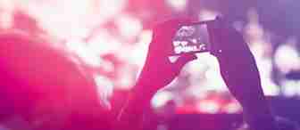 A person holding a smartphone in the air, recording or taking a photo at a concert. The background is filled with bright, colorful lights and blurred crowd, highlighting the energetic atmosphere of the event.