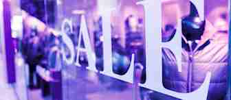 A store window with large white letters spelling "SALE" in a brightly lit retail environment. Inside, blurred mannequins display winter jackets. The scene has a purple-tinted lighting effect.