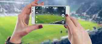 A person with red-painted nails is holding a smartphone, capturing a photo of a soccer field in a large stadium. The stands are filled with spectators, and teams are playing on the brightly lit field.