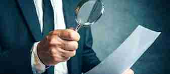 A person wearing a suit and tie is holding a magnifying glass in their right hand and examining a sheet of paper in their left hand. The background is blurred and has a dark, textured appearance.