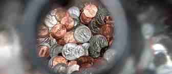 Close-up view of a mix of U.S. pennies, nickels, quarters, and dimes inside a jar, with the image being captured through the jar's opening, creating a vignette effect around the edges.