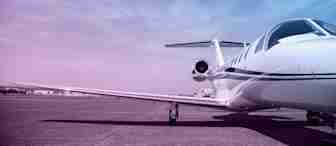 A sleek private jet is parked on an open tarmac under a clear sky with a gradient of purple and blue hues. The jet's wings extend outward, and the view focuses on the front portion and one of the engines. The landscape is open and expansive.