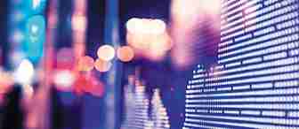 A close-up of a digital screen displaying financial data with charts and graphs made up of small illuminated dots. The background shows an out-of-focus view of bright city lights, creating a colorful bokeh effect. The scene has a modern and dynamic feel.