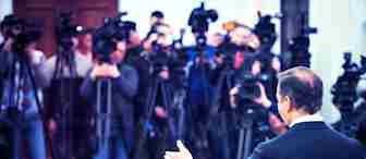 A person in a suit is speaking at a press conference. They are facing an audience filled with multiple cameras and reporters. The background is crowded with photographers capturing the event, and the room appears busy and active.