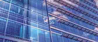A modern skyscraper with a glass facade reflects the sky, clouds, and neighboring buildings. The image is taken from a low angle, emphasizing the height and sleek design of the structure. The blue tones create a serene, contemporary atmosphere.