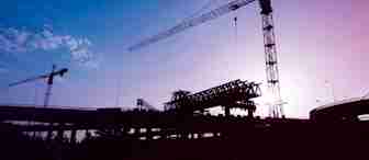 Silhouetted construction site with cranes working on a large structure against a purple and blue sky at twilight. The scene includes multiple cranes and the framework of a building or bridge with scattered clouds visible.