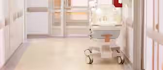 A hospital hallway with a neonatal incubator on wheels positioned near a closed door, under bright overhead lighting, and surrounded by white walls and medical equipment.