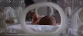 A newborn baby lies on its stomach inside a hospital incubator. The focus is on the baby's head, with soft lighting and a blurred background. The incubator's round openings surround the infant, providing a glimpse into the care setting.