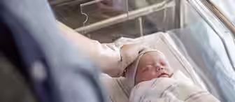 A newborn baby wrapped in a blanket sleeps in a hospital bassinet while an adult hand gently rests on the baby's head. The adult has a hospital ID band on their wrist.