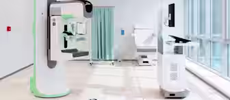 A modern, bright medical room with advanced diagnostic machines, a computer workstation, an examination chair, and a green privacy curtain near large windows letting in natural light.