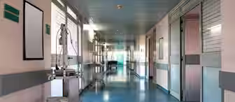 A long, empty hospital corridor with blue flooring and light-colored walls. Medical equipment is stationed on the left, and doors to patient rooms line both sides. Soft lighting illuminates the clean and orderly hallway, creating a calm atmosphere.