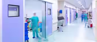 A bright hospital corridor with motion-blurred figures of medical staff in blue scrubs moving around. The hallway features sliding doors with visible medical equipment and neatly organized bins on the side.