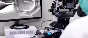 A laboratory technician wearing a green cap and white coat is using a microscope connected to a computer monitor displaying a magnified image of an egg cell undergoing a medical procedure, possibly in-vitro fertilization (IVF) or microinjection.