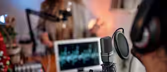 A close-up of a podcasting setup showing a microphone in the foreground, with two people talking in the blurred background. A laptop displaying an audio waveform and a sound mixer are on the desk, along with a turquoise mug.