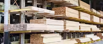 Stacks of neatly arranged wooden planks and beams are stored on metal racks in an outdoor lumber yard. The planks vary in size and thickness, and the storage area is covered by a roof.