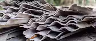 A close-up view of several stacked, weathered, and broken corrugated asbestos cement roofing sheets with a blurred outdoor background.