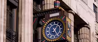A colorful, ornate clock with a blue face and gold hands is mounted on the exterior of a stone building with columns. The clock features geometric, multicolored patterns and stands out against the building's neutral facade.