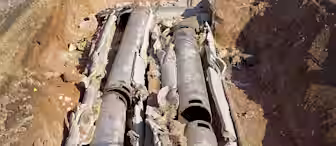 Three large, weathered metal pipes are laid side-by-side in a trench. The pipes are surrounded by insulation materials and dirt. The trench walls are made of uneven soil with exposed edges. The overall scene suggests ongoing construction or maintenance work.