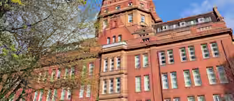 A large red-brick building with multiple windows and a distinctive central tower stands against a bright blue sky. Trees with budding leaves frame the left side of the image. The architecture is classic and intricate, indicating an older, possibly institutional building.