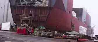 Partially constructed ship with exposed metal framework and scaffolding, surrounded by construction equipment and materials. The background is foggy, and the environment suggests a shipyard or industrial setting.