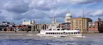 Image of a river scene with a large white boat cruising along. The background features a mix of modern and historical buildings, including a prominent domed structure under a partly cloudy sky. There is a sense of movement in the water and the cityscape appears lively.