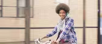 A person with a big afro hairstyle, wearing a plaid shirt, patterned scarf, and headphones, is smiling while riding a green bicycle. The background features a fence and a building.