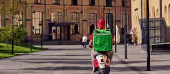 A food delivery person wearing a red cap and shirt rides a scooter with a large green insulated bag on their back through a modern urban courtyard with brick buildings.