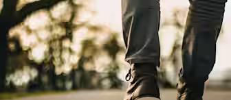 Close-up of a person walking on an outdoor path, wearing dark brown leather boots and gray pants. The background is softly blurred, featuring trees with a warm, golden light suggesting a sunset or sunrise.