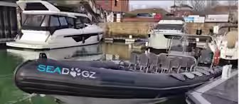 A black inflatable boat, labeled "SEADOGZ," is docked in a marina. It's empty, with multiple rows of gray seats. Other boats and a brick building are visible in the background. The water is a greenish hue.