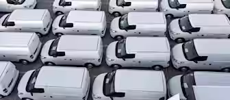 Aerial view of rows of white delivery vans parked in a lot. The vehicles are closely spaced, aligned in a neat grid pattern, reflecting their roofs and side panels under the light.