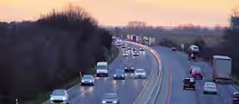 A highway at sunset with a mix of cars and trucks traveling in both directions. The left side of the road is busier with several vehicles, while the right side has fewer vehicles. Trees border the highway on the left, and open fields are visible on the right.