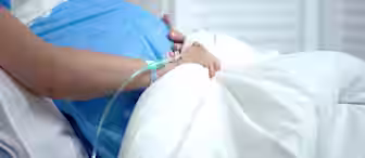 A pregnant person lying in a hospital bed, wearing a blue gown. Their hand is resting on their belly and has an IV line attached. The background consists of white curtains, and the scene suggests a medical or hospital setting.