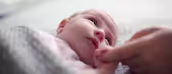 A baby lies on a soft, grey blanket, looking up. The baby is wrapped up in the blanket, and a person’s hand is gently holding the baby’s hand. The scene evokes a sense of warmth and tenderness.