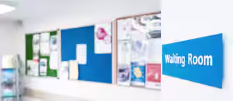 A hospital or clinic waiting room with light blue chairs and a notice board filled with various flyers and notices. A blue sign on the wall reads "Waiting Room.