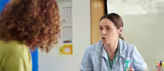 A nurse in scrubs sits at a desk, speaking with a patient. The nurse gestures with her hands, while the patient listens. Medical equipment and paperwork are visible on the desk.