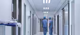 Two medical professionals in blue scrubs and caps walk down a well-lit hospital corridor. The hallway is lined with doors and a medical cart is visible on the left side. The setting is clean and sterile.