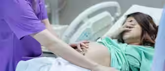 A pregnant woman lies in a hospital bed with her eyes covered. A medical professional in a purple uniform examines her belly. Another person stands nearby. The setting is a medical facility, likely a maternity ward.