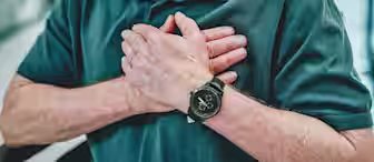 A person wearing a dark polo shirt and a watch holds their chest with both hands, indicating possible chest pain or discomfort. The background is blurry but appears to be indoors.