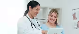 A doctor in a white coat shows information on a tablet to a smiling pregnant woman sitting beside her in a medical exam room.