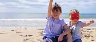 Two young children sit on a sandy beach by the ocean under a blue sky. One child has short brown hair, wearing a blue shirt and denim shorts, and raises an arm in the air. The other child wears a superhero mask and also raises an arm, mimicking flying.