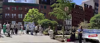 A group of people stands in a sunny plaza, attentively listening to a woman speaking at a microphone. A large banner reading "ACTION MESOTHELIOMA" is displayed to the right. Surrounding buildings and a few trees are visible in the background.