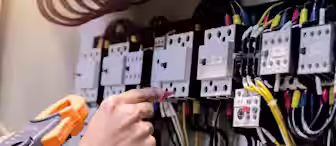 A person uses a handheld clamp meter to measure electrical current in an industrial electrical panel. Multiple wires and circuit components are visible, with the person pointing to one of the components. The setting appears to be a maintenance or technical environment.