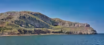 A coastal cliff covered in greenery rises against a clear blue sky, with the rocky formations extending into a calm blue sea. The landscape features a mix of rocky ledges and grass patches, showcasing natural beauty.