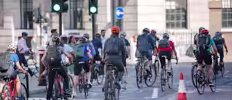 A group of cyclists, all wearing helmets and backpacks, ride through an urban intersection. The street features a traffic light, and there's a traffic cone placed nearby. The background shows a building with large windows and pedestrians walking along the sidewalk.