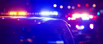 A night scene featuring several police vehicles with flashing blue and red lights illuminating the dark surroundings. The focus is on the lights of the police cars, creating a sense of urgency or alert. The background is blurred with additional flashing lights visible.