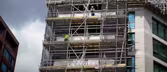 A construction site on a high-rise building covered in scaffolding. Several workers in safety gear are visible at different levels. The building appears to be under renovation or construction against a backdrop of a partly cloudy sky.
